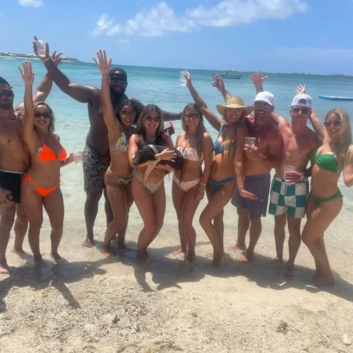 Cheerful group with drinks celebrating on a beach sandbar, one holding a pig, near Nassau, Bahamas