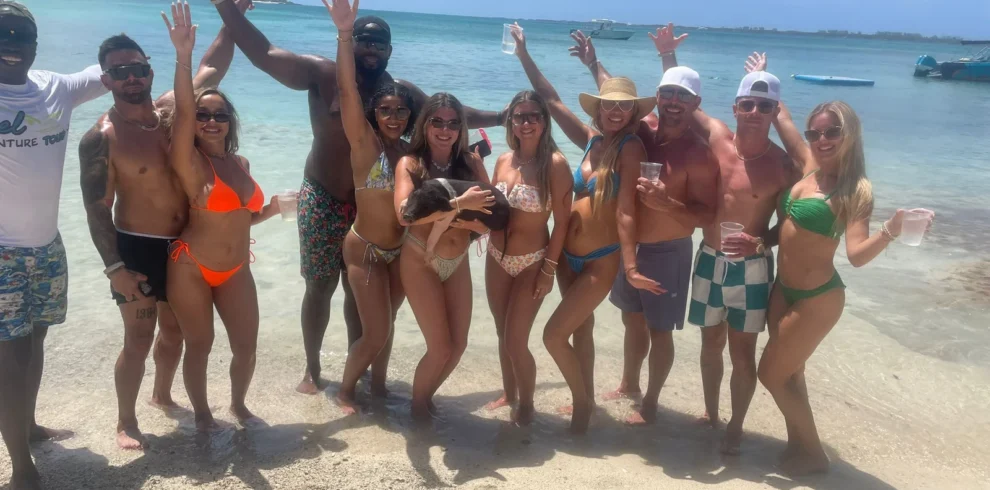 Cheerful group with drinks celebrating on a beach sandbar, one holding a pig, near Nassau, Bahamas